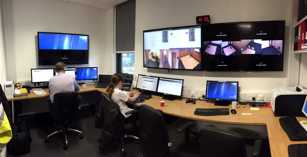 Two people operating a Hydra control desk beneath wall-mounted displays