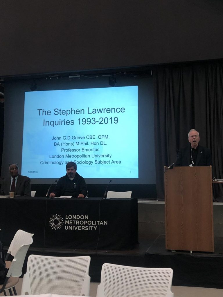 Speaker at a lectern beside a Stephen Lawrence Inquiry conference presentation